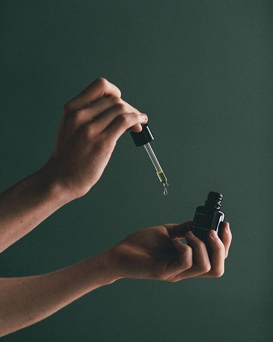 Hands holding a small dark glass dropper bottle of essential oil against deep green